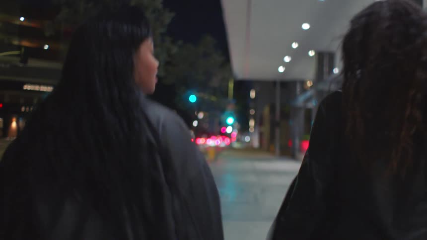 Two young women wearing black jackets and long boots walking along a city sidewalk at night illuminated by streetlights