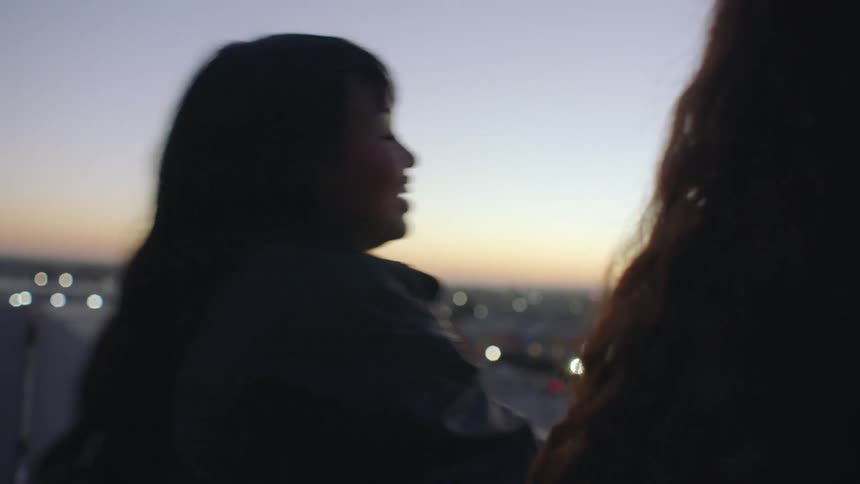 Two women stand together on a rooftop at twilight looking out over the city lights while talking and smiling as the sun sets in the background