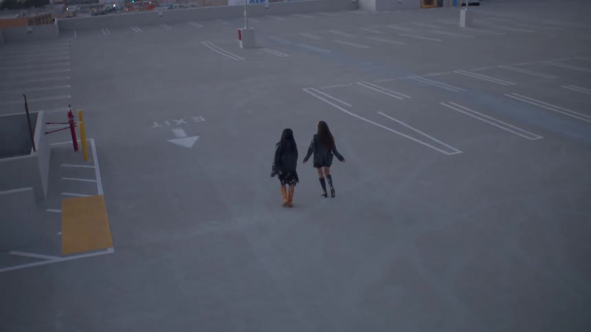 Two women walk across a sprawling empty rooftop parking lot toward a dense city skyline at dusk captured from a highangle perspective
