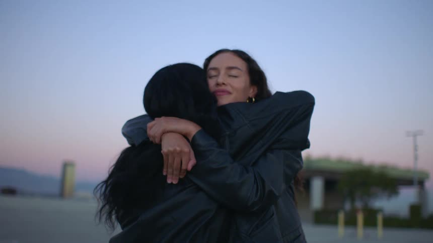Two women wearing black leather jackets share a long meaningful hug on a rooftop at twilight before pulling apart and smiling at each other with the city skyline and evening sky visible in the blurred background