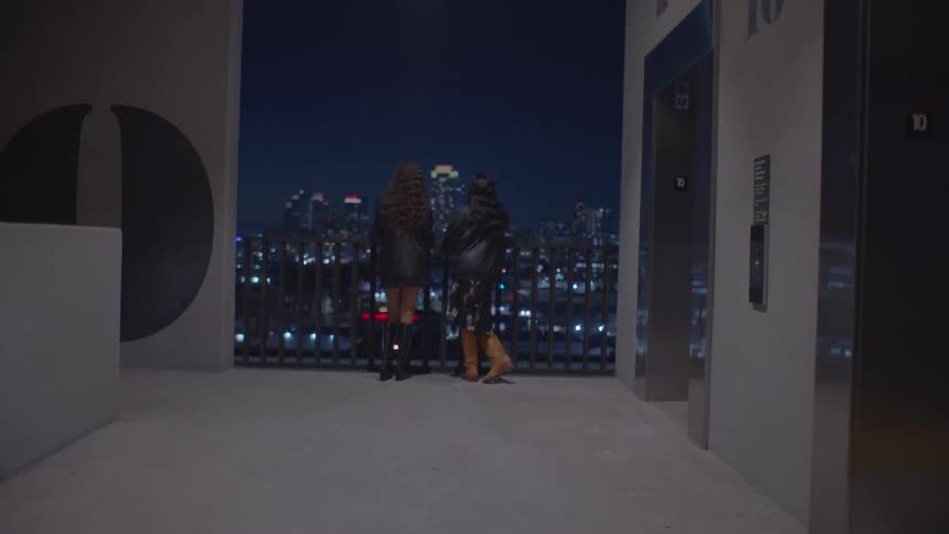 Two young women standing on a balcony at night looking out at the city skyline illuminated in the background