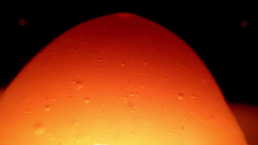 A glowing orange domeshaped mass of viscous liquid rises slowly against a dark background tiny bubbles are visible moving within the translucent substance as it gradually shifts and flows upward