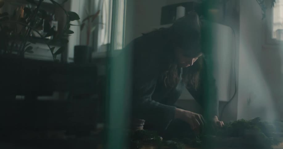 Woman seated indoors handling plants on a table near a window