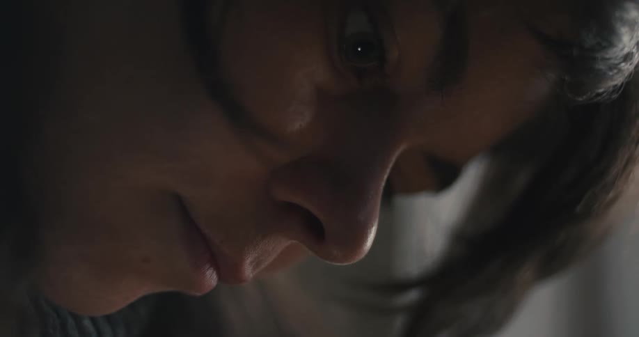 A dramatic closeup captures a womans face in soft dim light her eyes cast downward with an intense contemplative expression that emphasizes the subtle texture of her skin and the shadows along her nose and cheek
