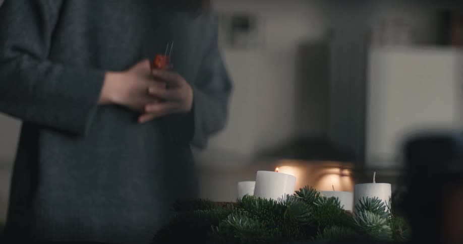 A woman in a grey sweater uses a lighter to ignite one of four white candles on a handmade evergreen wreath standing quietly in a dimly lit room with a thoughtful expression