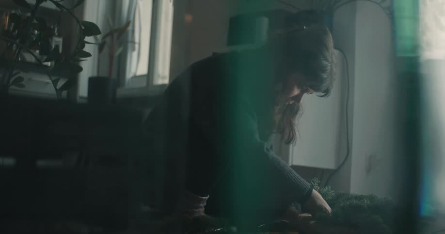 A woman kneels on the floor of a dimly lit room carefully assembling a green holiday wreath from pine branches under a soft light that creates a prism effect across the frame