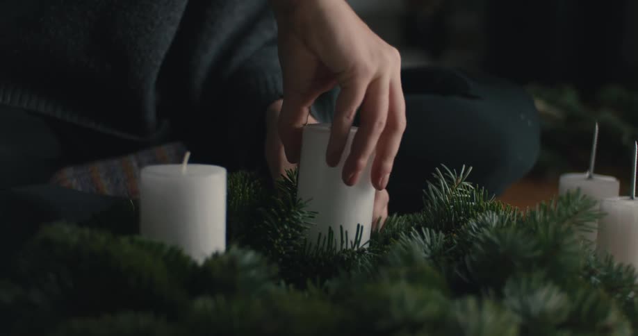 A woman with long dark hair wearing a gray sweater sits on the floor and carefully nestles a thick white candle into a handmade evergreen wreath