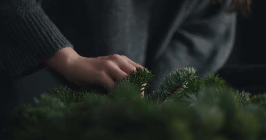 A closeup shot captures the steady hands of a woman wearing a dark knit sweater as she arranges a lush circular evergreen wreat
