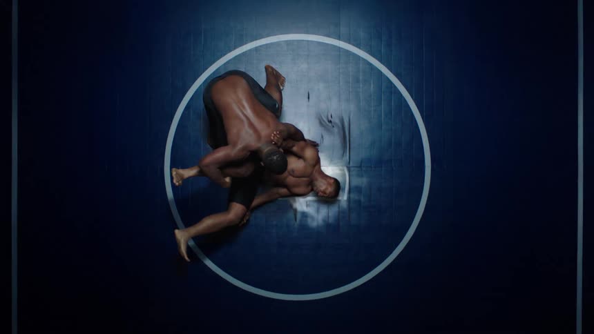 Two male wrestlers are grappling on a dark blue mat locked together inside a white circle in the center of the ring