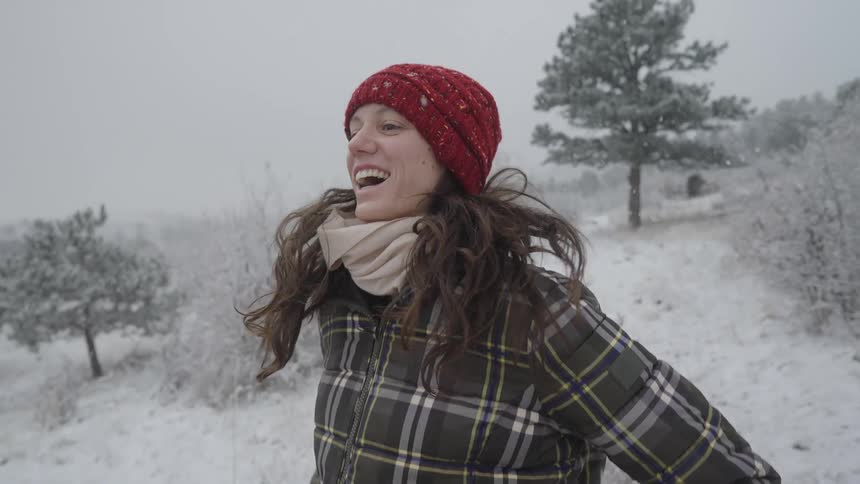 A woman wearing a red knit hat plaid winter coat scarf and gloves smiles as she jumps outdoors in a snowy landscape with trees in the background