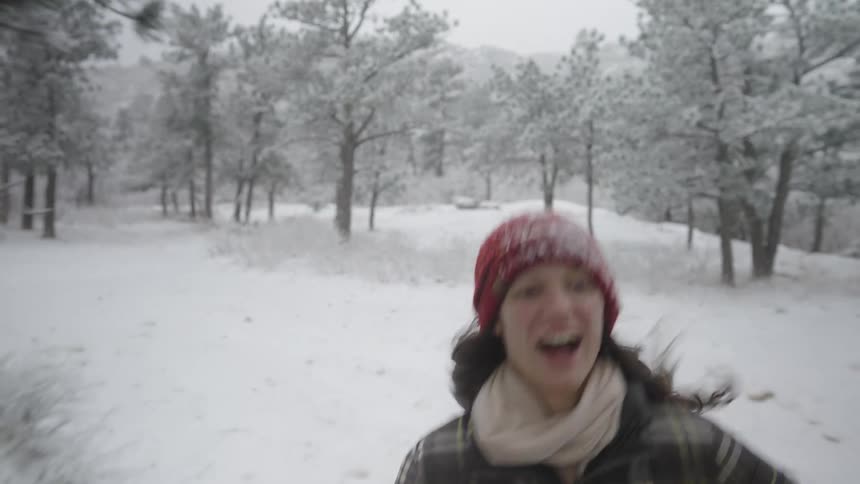 A woman with long dark hair wearing a plaid jacket and a red knit hat runs toward the camera and laughs while playing in a snowy forest