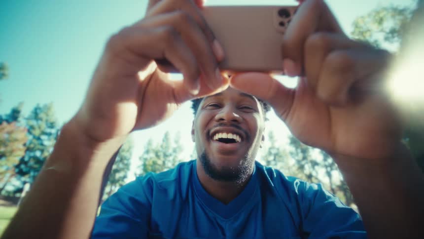 The face of a smiling man who is fiddling with a cell phone while sitting at a table in a grassy yard
