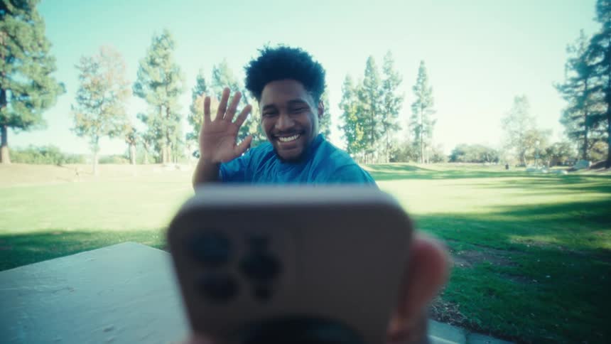 A black man takes a selfie with his smartphone while sitting at a picnic table in a park