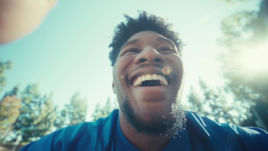 A man in a blue shirt holds a smartphone close to his face outdoors while sitting at a bench looking up at the screen with a surprised expression as sunlight shines behind him