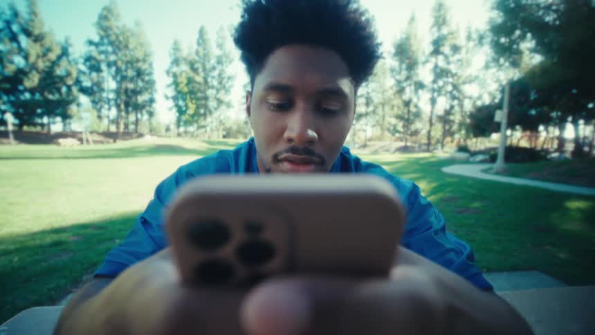A black man sits at a park on a table using and reading on a smart phone
