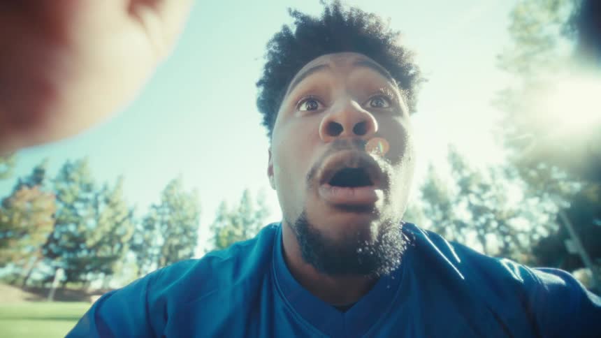 A man in a blue jersey sits at a table in a sunlit park and holds a smartphone horizontally to record his face transitioning from focused to wideeyed shock as the camera zooms in