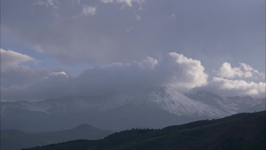Thick white clouds drift quickly across the snowy peaks of a massive mountain range under a vast sky while dark green foothills sit quietly in the foreground during this scenic daytime timelapse