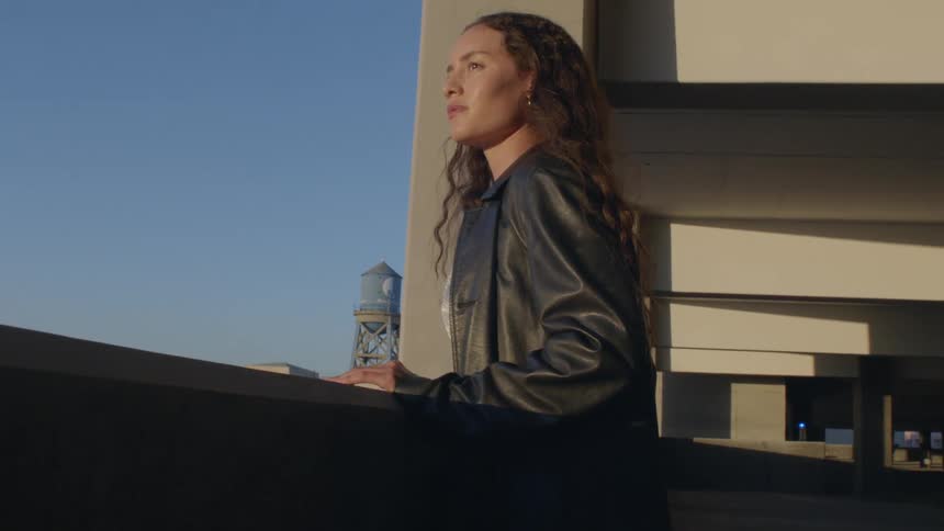 Young adult woman wearing a leather jacket looking at an urban area from the balcony of a parking lot building outdoors during day time