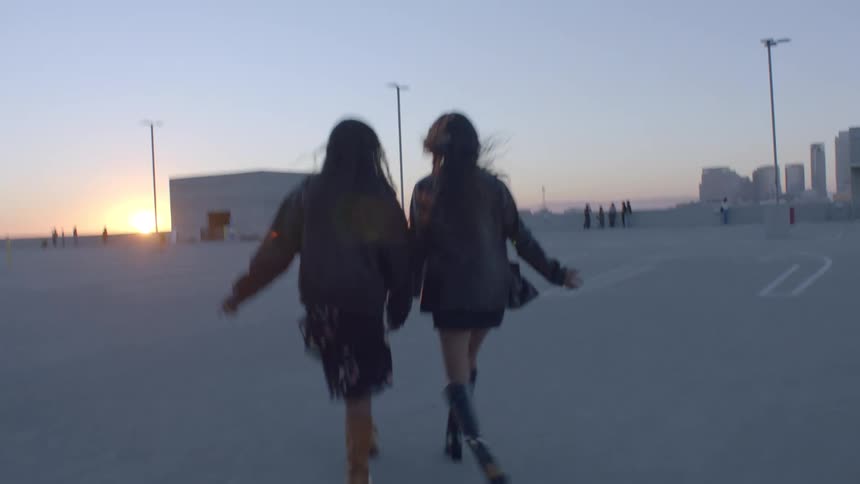 Two women walk together across an empty rooftop parking lot at sunset