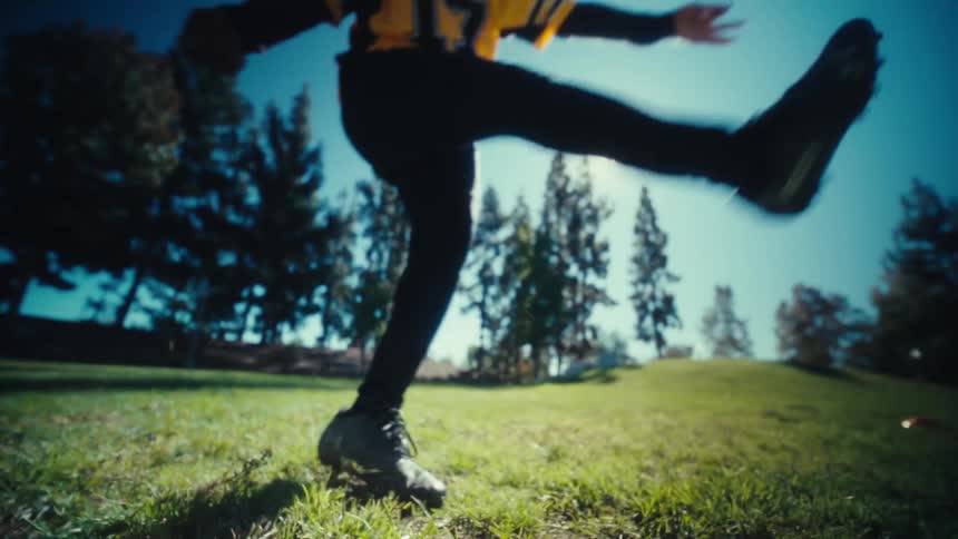 From a low angle a boy in a yellow jersey runs into frame and powerfully kicks a football off a tee sending it soaring into the bright sunlight