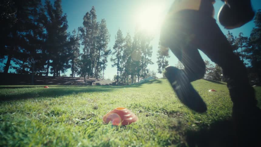A black man practices football by running around orange cones in a sunny grassland