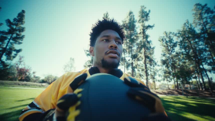 A man in a yellow football jersey stands in a sunny park holding a ball and looking toward the horizon while the camera captures him from a low angle