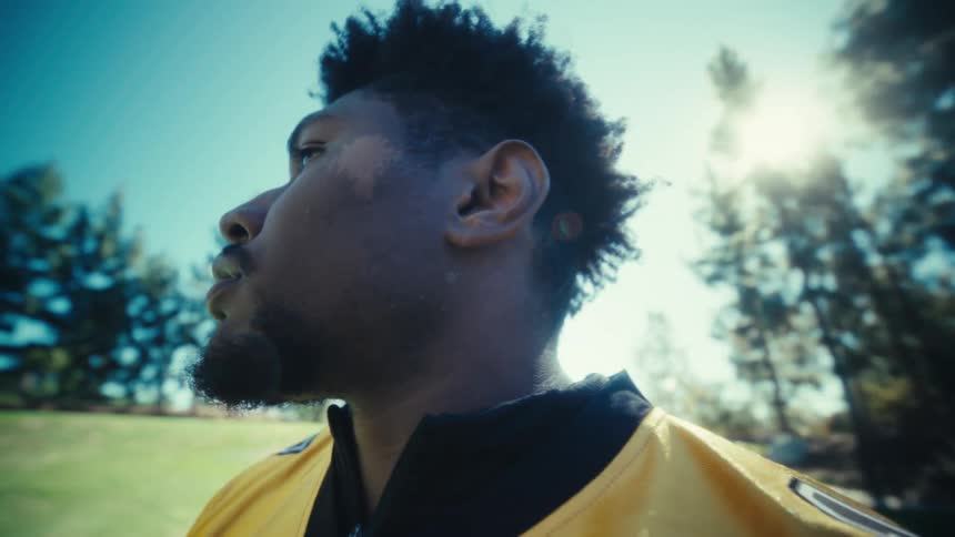 An american football player in a yellow jersey stands on a grassy field while looking around the park holding some equipment as the camera follows him under a bright sun