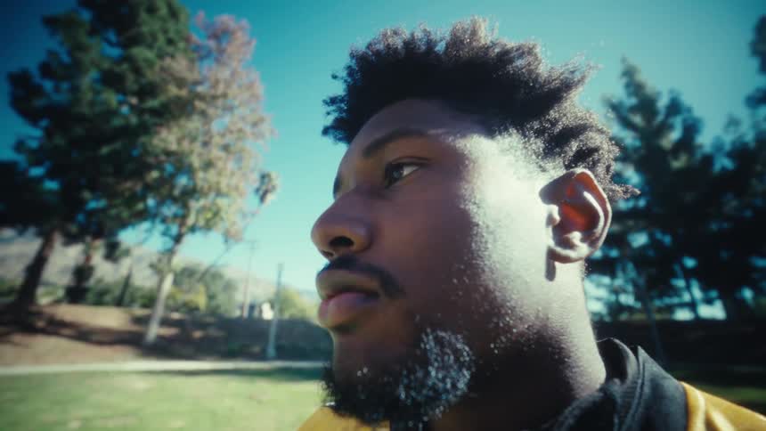 A man with a beard and short afro stands in an outdoor park captured in a lowangle closeup as he looks off into the distance under a bright sun