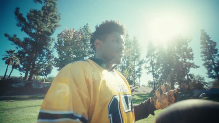A man wearing a yellow and black football jersey with the number seventeen stands on a green field holding a football and looking toward the camera on a bright sunny day with tall trees in the background