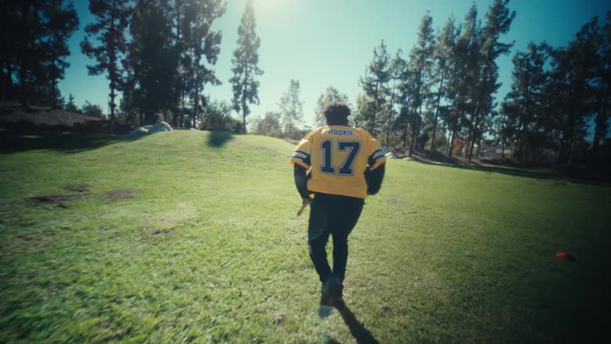 A football player in a yellow and black jersey runs through agility cones on a sunlit grass field the camera follows him closely as he sprints weaves and demonstrates athletic footwork while clutching a football