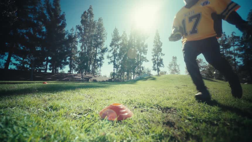 A man wearing a yellow football jersey runs across a green grass field toward an orange marker during a daytime practice session with tall trees and bright sun in the background