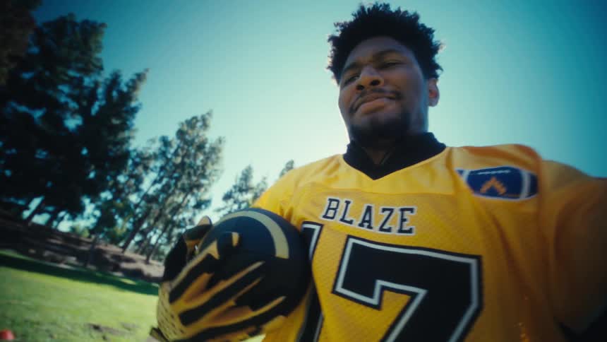Young black male running through a public park on a sunny day holding a football and wearing a yellow jersey
