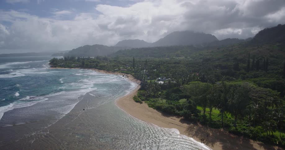 A curving tropical coastline where waves roll onto a wide sandy beach bordered by lush green vegetation with misty cloudcovered mountains rising in the background under a dramatic sky