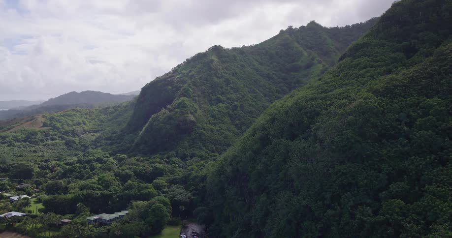 An aerial perspective glides over steep lush mountains covered in dense tropical greenery while a small parking lot and buildings sit nestled in the valley below