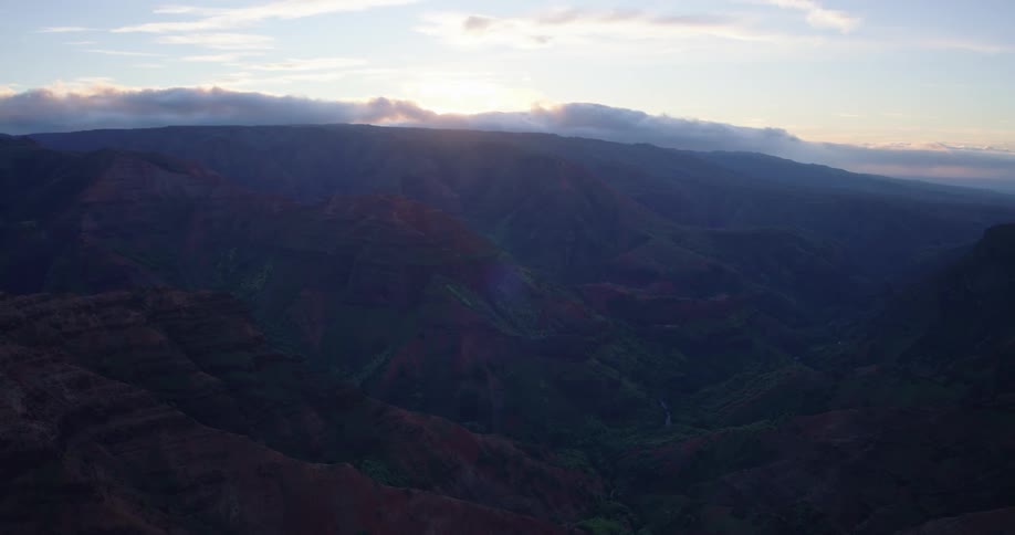 The sun sets behind a line of low clouds above a vast rugged canyon landscape casting soft light and shadows over the layered red and green slopes that stretch into the hazy distance