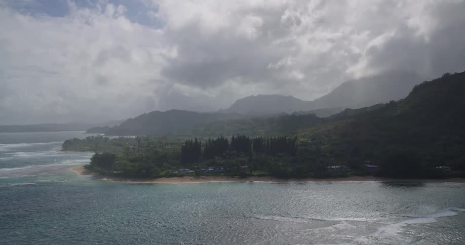 An aerial camera banks over a turquoise coastline and breaking waves before moving toward a misty green mountain range under a heavy overcast sky