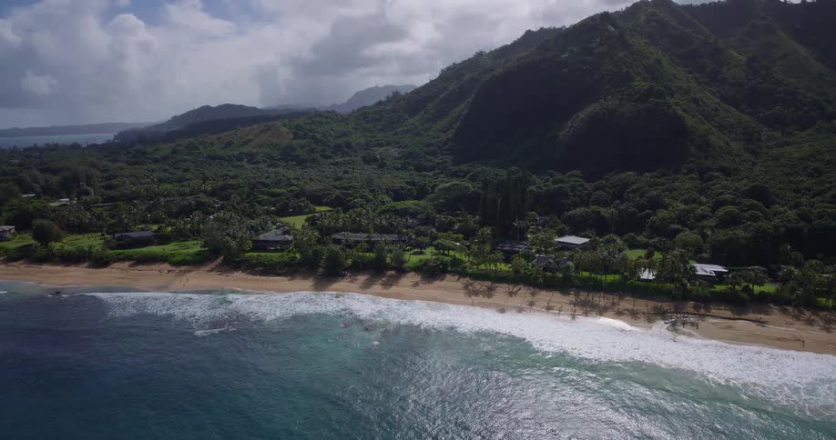 Aerial view of mountains and homes along a lush coastline with waves rolling onto a sandy beach