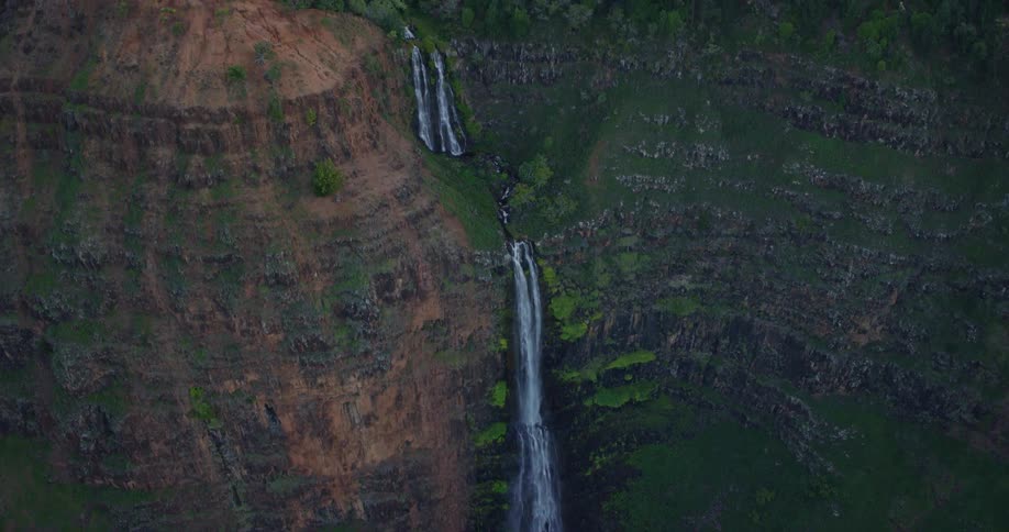An aerial drone shot reveals a narrow waterfall cascading down a sheer multicolored cliff face into a deep canyon the camera tilts upward to show the lush green forest at the top of the plateau under soft overcast lighting