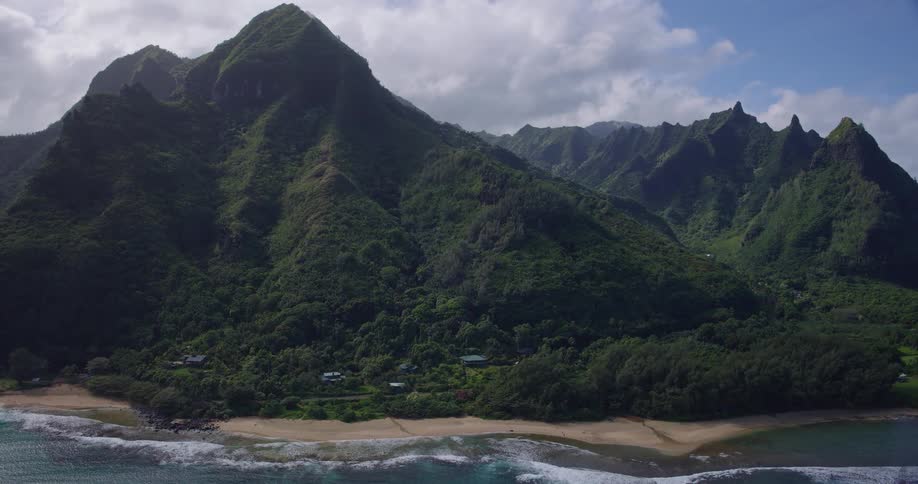 Steep mountains covered in vegetation surround beach with gentle waves and buildings among trees