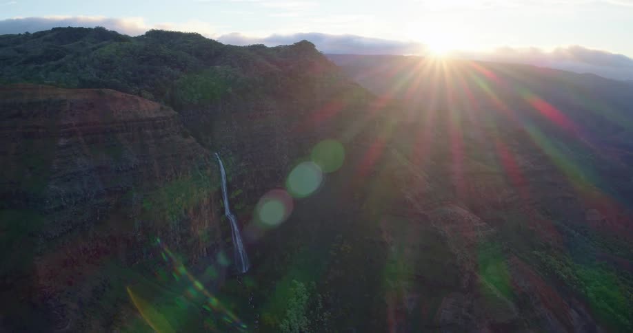 A tall waterfall cascading down a cliff face in a vast verdant canyon as the sun flares brightly over the mountainous horizon