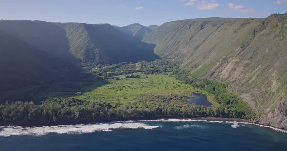 An aerial view of a secluded coastal valley shows steep towering green cliffs enclosing a broad grassy plain with scattered trees and a small lake bordered by a narrow strip of forest and white waves breaking along the deep blue shoreline