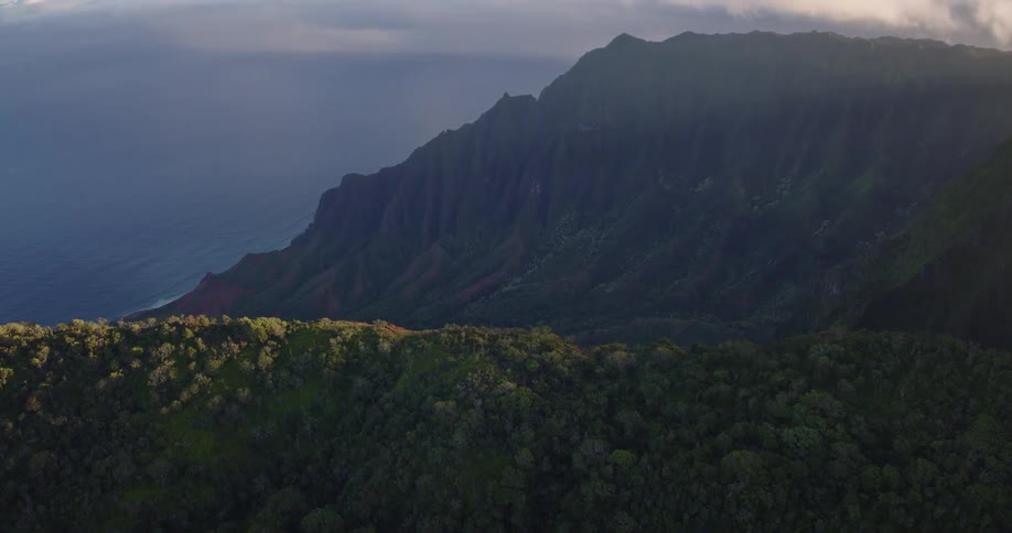 An aerial camera rises from a densely forested ridge to reveal massive jagged cliffs along a tropical coastline as golden light strikes the peaks under a cloudy sky