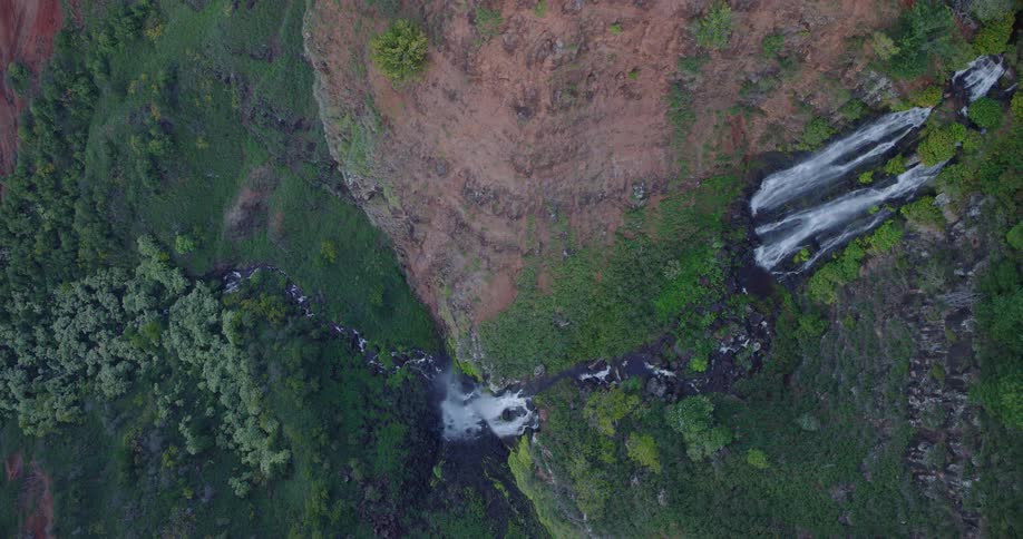 An aerial camera glides forward and upward over a sheer rocky cliff trailing the descent of multiple waterfalls into a lush tropical canyon under soft natural daylight