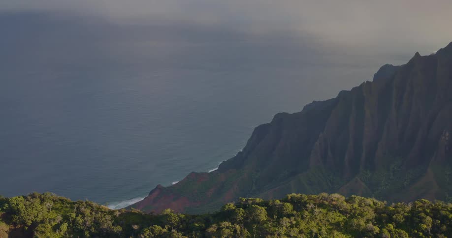 Aerial view of a coastal mountain range with sharp green ridges meeting a deep blue ocean under a hazy sky at sunset
