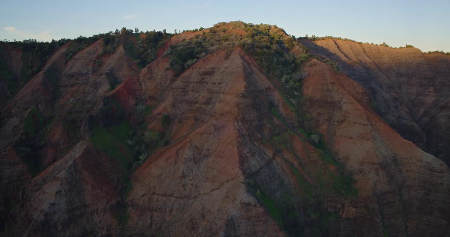 An aerial shot of a deep green valley surrounded by steep cliffs that open up to a dark sand beach and blue ocean the second video shows the rugged multicolored ridges of a vast canyon illuminated by the warm light of a setting sun