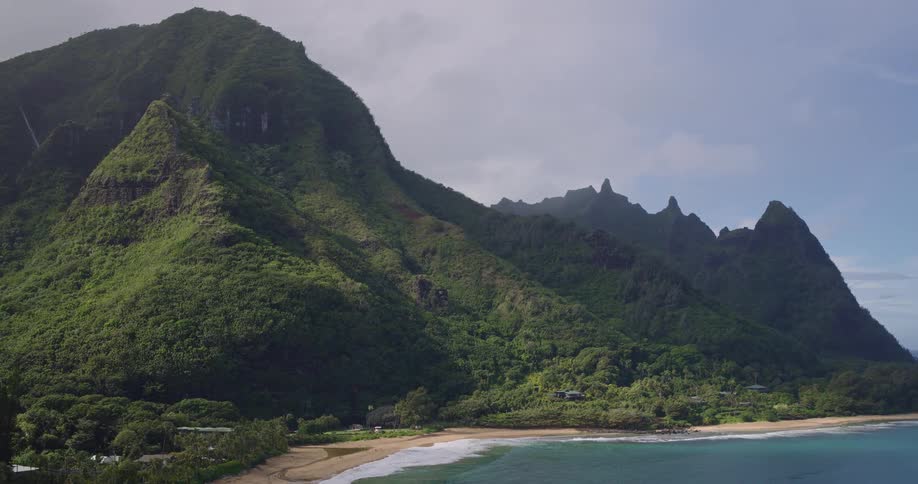 An aerial view captures the stunning tropical coastline where lush green mountains meet a serene sandy beach and the gentle ocean waves