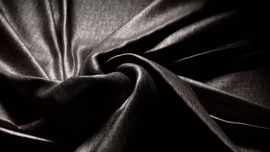 Starting as a flat surface dark textured fabric is slowly pulled and twisted into a central spiral under a focused light creating deep rhythmic folds and shifting highlights as it rotates into a vortex within the darkness