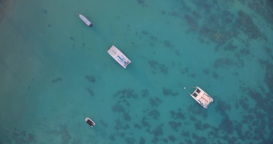 Four boats are anchored in clear turquoise water revealing shadowy reef or seabed patterns below