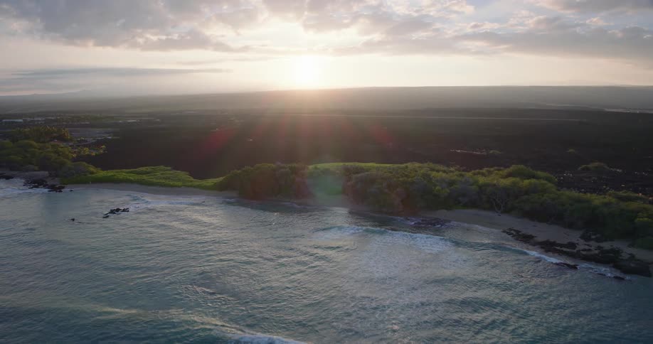 An aerial camera glides backward over gentle ocean waves as the sun sets over a rugged coastline the golden light illuminates a lush green shoreline contrasted against a vast dark volcanic landscape