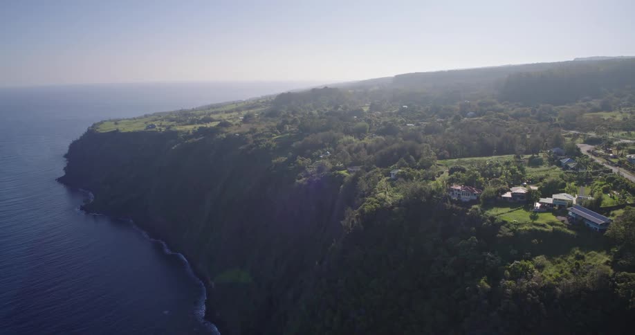 Aerial view of a high green cliff overlooking the deep blue ocean with houses and a winding road nestled among lush trees under a clear bright sky